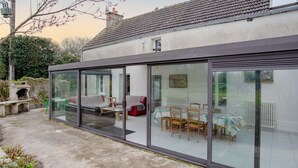 A modern house with a glass-enclosed patio, a dining area with wooden chairs, and a red sofa. - Les Mésanges by Interhome (St. Lo-d'Ourville)