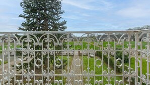 Un balcón con barandillas de metal blanco ornamentado que da a un césped cuidado y un alto árbol perenne.