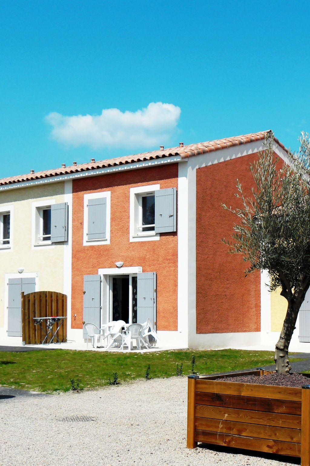 A two-story building with a red brick facade, white trim, and a tiled roof. There is a small patio with outdoor furniture and a wooden gate.