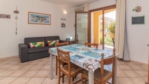 A dining area with a table covered in a patterned tablecloth, surrounded by wooden chairs. A gray sofa with colorful cushions is against the wall, and a painting is hung above it. - Alessandra by Interhome (Santa Teresa di Gallura)