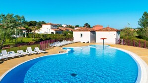 A large, clear swimming pool with lounge chairs and a red umbrella, surrounded by a fenced area and buildings in the background. - Port Minervois by Interhome (Homps)