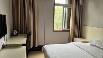 A hotel room with a bed, a desk with a chair, a window with curtains, and a television on the wall.