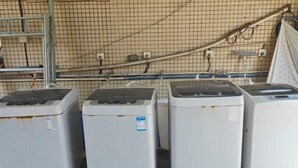 Quatre machines à laver de style industriel sont alignées contre un mur, sous un plafond à motifs en forme de grille, et à proximité de prises électriques.