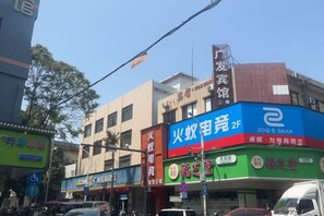 A multi-story building with commercial establishments on the ground floor, including a shop with a blue sign and another with a red sign. - Guangfa Hostel (Guangzhou Nansha) (Guangzhou)
