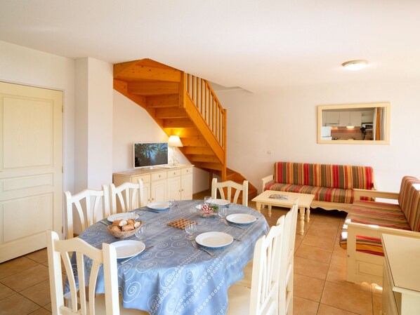 Un coin repas avec une table dressée pour quatre personnes, un escalier en bois, un canapé et une cuisine visible au fond.