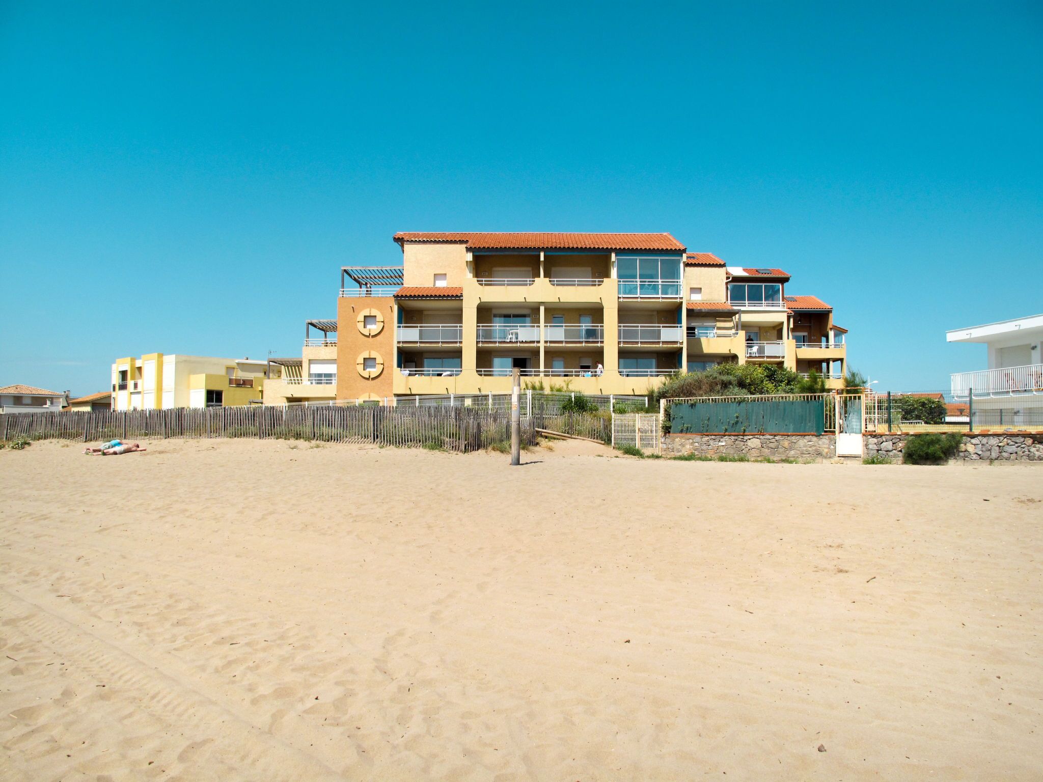 Un edificio a più piani con balconi, una spiaggia sabbiosa in primo piano e un cielo azzurro limpido.