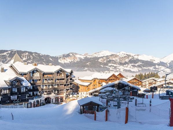 A snow-covered mountain resort with wooden buildings, ski lifts, and a clear view of snow-capped mountains in the background.
