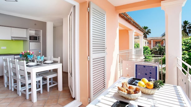A modern kitchen with white cabinets and a green countertop, a dining area with white chairs and a table, and a balcony with a table set with fruits and pastries. - Résidence Odalys Référence La Palmeraie by Interhome (Grimaud)
