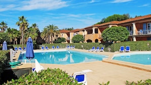 A pool area with multiple lounge chairs and umbrellas, surrounded by palm trees and a building with a tiled roof. - Résidence Odalys Référence La Palmeraie by Interhome (Grimaud)