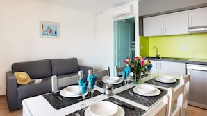 A dining area with a table set for four, a grey sofa, a kitchen with a green backsplash, and a door leading to another room. - Résidence Odalys Référence La Palmeraie by Interhome (Grimaud)