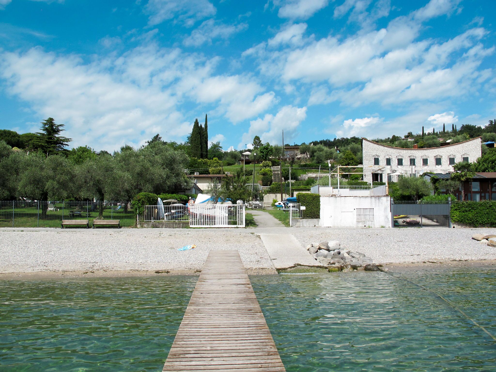 A wooden pier leading to a calm body of water, surrounded by a fenced park area with benches and a white building in the background.
