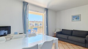 A modern living room with a flat-screen TV, a grey sofa, a dining table with chairs, and a large window with curtains. - Résidence Odalys Référence La Palmeraie by Interhome (Grimaud)
