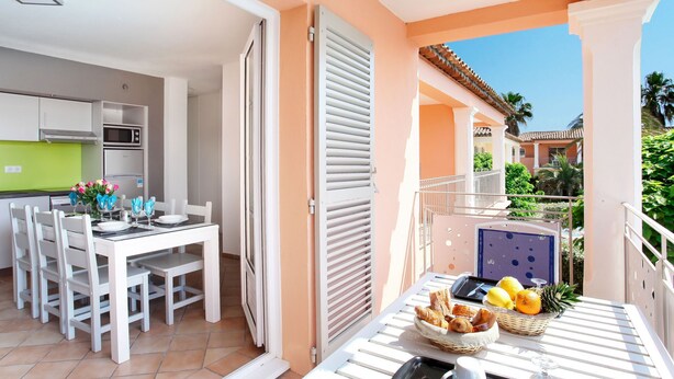 A modern kitchen with white cabinets and a green countertop, a dining area with white chairs and a table, and a balcony with a table set with fruits and pastries. - Résidence Odalys Référence La Palmeraie by Interhome (Grimaud)