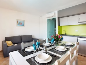 A dining area with a table set for four, a grey sofa, a kitchen with a green backsplash, and a door leading to another room.