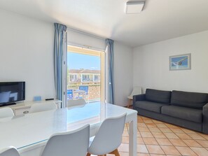A modern living room with a flat-screen TV, a grey sofa, a dining table with chairs, and a large window with curtains.
