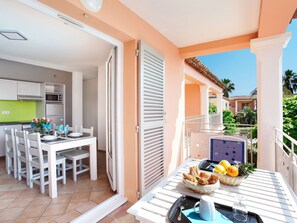 Una cocina moderna con gabinetes blancos y una encimera verde, una zona de comedor con sillas blancas y una mesa, y un balcón con una mesa servida con frutas y pasteles.