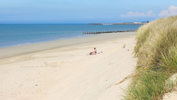 Une plage avec une personne assise sur le sable, une dune herbeuse et une mer calme.