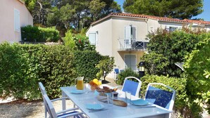 Un área de comedor al aire libre con una mesa lista para dos personas, rodeada de vegetación y un edificio con balcón.