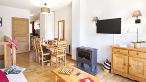 Un salon chaleureux avec une table à manger en bois, des chaises, une cheminée, un téléviseur fixé au mur et un buffet en bois.