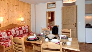 A cozy room with a wooden wall, a sofa with a red and white patterned cover, a dining table set for four, and a kitchen area with a refrigerator. - Odalys Référence Le Grand Panorama 1 by Interhome (Valmeinier)