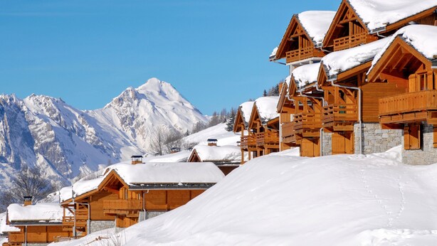 A row of wooden chalets with snow-covered roofs, set against a backdrop of snow-capped mountains under a clear blue sky. - Odalys Référence Le Grand Panorama 1 by Interhome (Valmeinier)