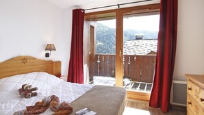 A bedroom with a bed, a teddy bear, a book, and a view of a balcony with red curtains. - Odalys Référence Le Grand Panorama 1 by Interhome (Valmeinier)