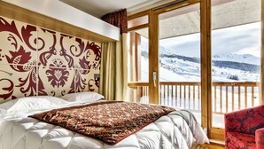 A hotel room with a large bed, a wooden headboard with a floral pattern, a red and beige patterned throw, and a view of a snowy landscape through a sliding glass door. - Résidence Odalys Collection Edenarc by Interhome (Les Arcs)