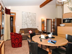 A modern living room with a round dining table, a sofa, and a red patterned armchair. There is a large wall art piece above the sofa and a kitchen area with a microwave and oven.