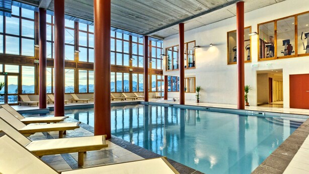 Une piscine intérieure dotée de chaises longues, de grandes fenêtres et offrant une vue sur les montagnes.