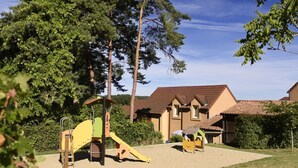 A playground with climbing structures and slides, surrounded by trees and a building with a tiled roof.