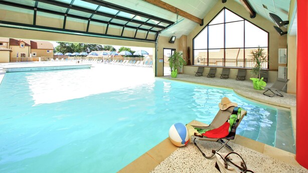 An indoor swimming pool with clear blue water, a lounge area with sunbeds, and a view of the outside buildings.