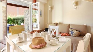 A bright room with a glass table set for breakfast, a sofa, and a view of greenery outside.