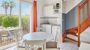 A compact kitchen with white cabinets, a microwave, and a sink. There is a dining area with a round table and chairs, and a wooden chair near a staircase. - Odalys Essentielle Le Grand Bleu by Interhome (Valras Plage)