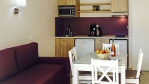 Un angolo cottura compatto con forno a microonde, macchina del caffè e un piccolo frigorifero. C'è un tavolo da pranzo con sedie e un cesto di frutta. Un divano rosso è posizionato di fronte all'angolo cottura.