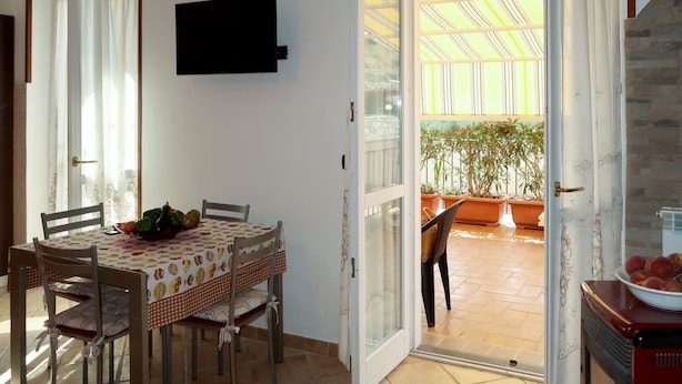 A dining area with a table set for four, chairs, a television mounted on the wall, and a view of a patio with potted plants. - Samuele by Interhome (Civezza)