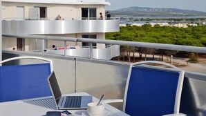 A balcony with a table, chairs, and a laptop. There is a view of a building and trees in the background. - Odalys Référence Le Lotus Blanc by Interhome (Le Barcarès)