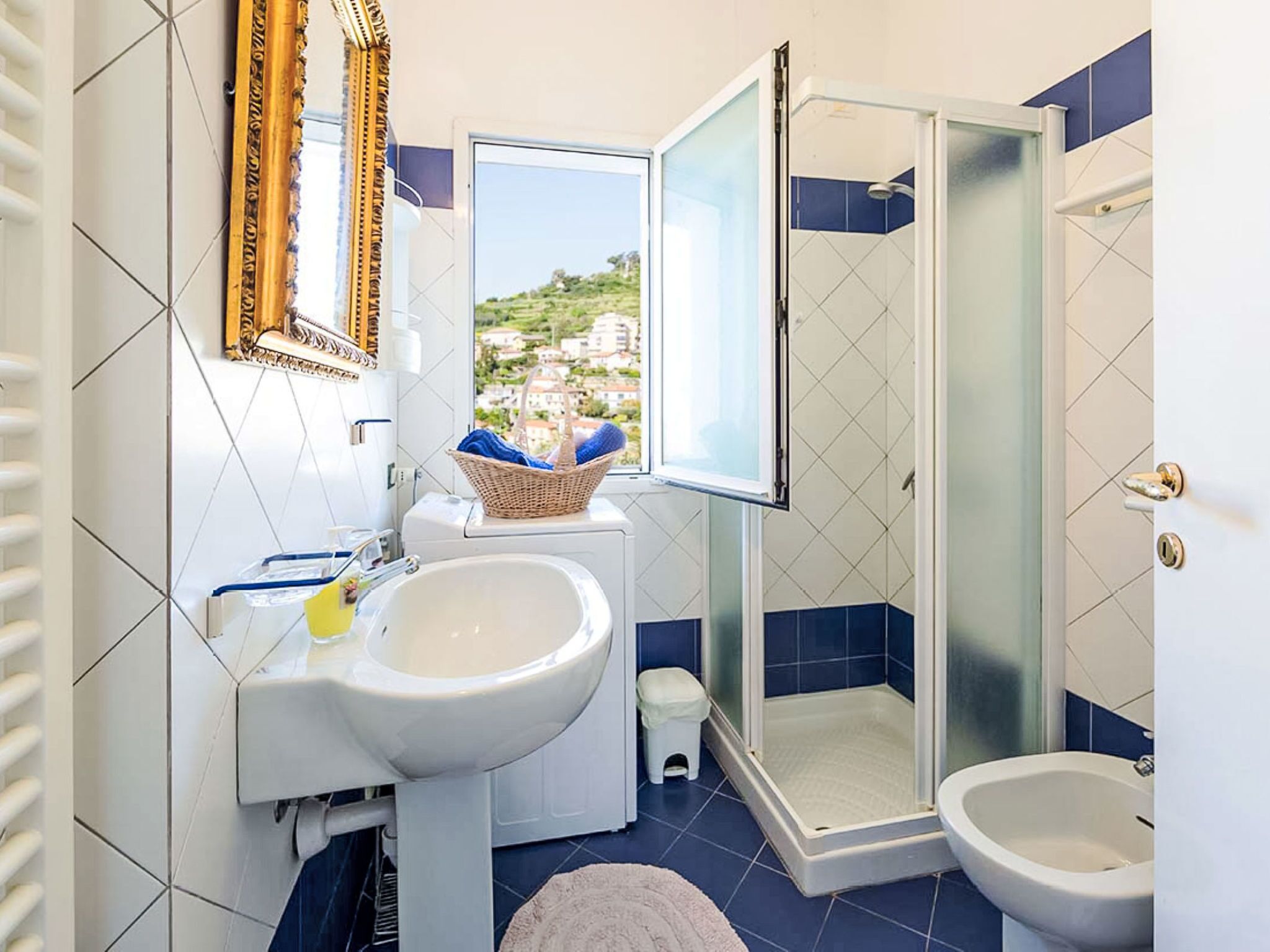 A bathroom with a sink, a bidet, a shower, and a window with a view of houses.