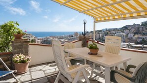 Une terrasse dotée d’un auvent à rayures jaunes, de chaises blanches et d’une table. On y trouve des plantes en pot et une vue sur la ville et la mer.