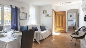 Une pièce comprenant une table à manger dressée pour deux personnes, un lit recouvert d’une courtepointe blanche, une armoire en bois et une chaise avec un coussin.