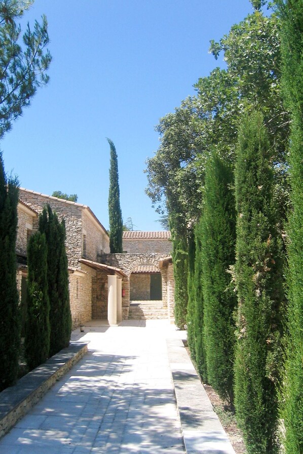 A stone building with a tiled roof, a paved pathway, and neatly trimmed evergreen trees.