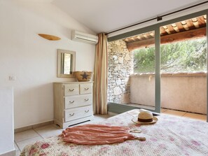 Quarto com parede de pedra, teto de madeira, cômoda branca, cama com colcha florida e janela com cortinas.
