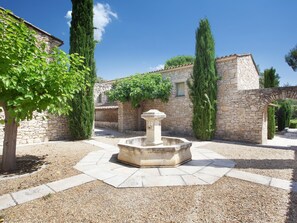 A courtyard with a stone fountain, surrounded by stone walls and tall cypress trees.