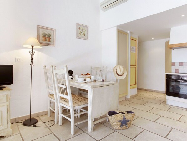 A dining area with a table set for two, a television on a stand, and a floor lamp. There are two chairs, a basket, and a hat on the wall.