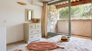 A bedroom with a stone wall, a wooden ceiling, a white dresser, a bed with a floral bedspread, and a window with curtains.