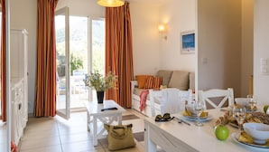 A bright and airy living room with a sofa, a dining table set with food and drinks, and a view of the outdoors through large windows. - Odalys Référence Les Villas Bel Godère by Interhome (Lozari)