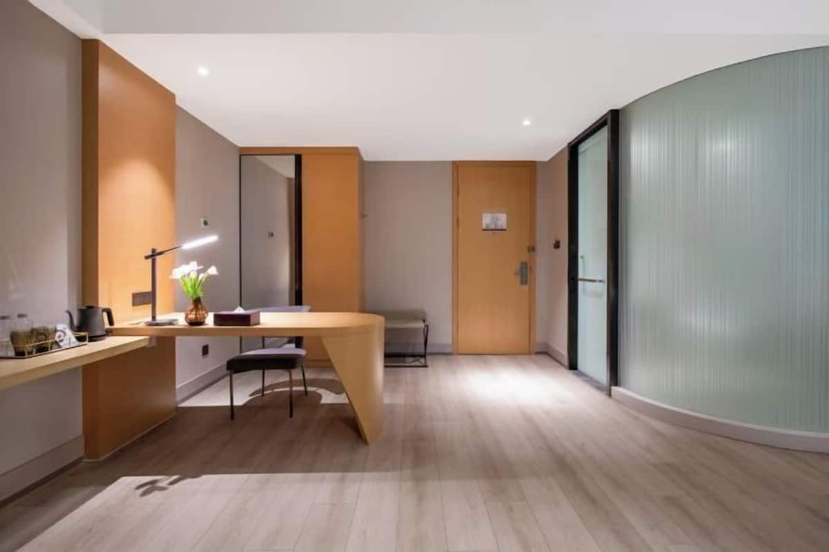 Una habitación de hotel moderna con un escritorio de madera, una silla, una mesita, un espejo y un separador de vidrio.