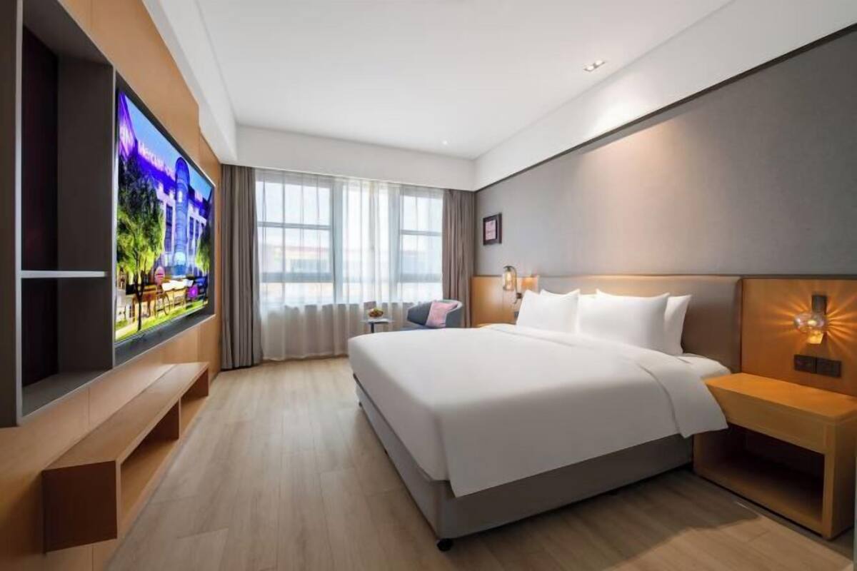 Una habitación de hotel moderna con una cama grande, un televisor montado en la pared y un ventanal con cortinas.