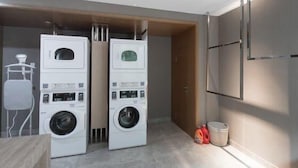 A laundry room with two stacked front-loading washing machines, a small trash bin, and a wooden door. - Mercure Qidong Downtown (QIDONG)