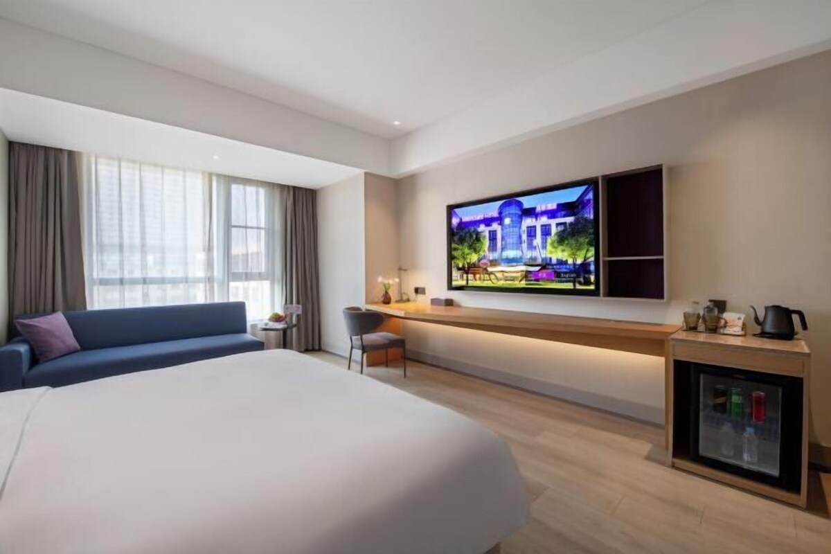 A modern hotel room with a large bed, a TV mounted on the wall, a small desk with a lamp, and a minibar.