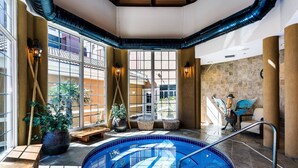 An indoor swimming pool with clear glass walls, wooden benches, and potted plants.